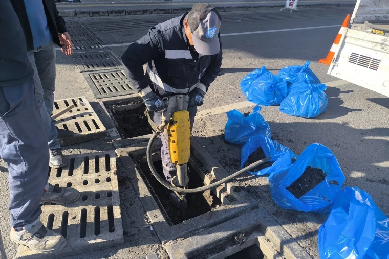 DİSKİ ekibi Diyarbakır'da bir caddede yağmur suyu ızgarasının içini kırıcıyla temizliyor, çıkan tortu mavi çuvallarda